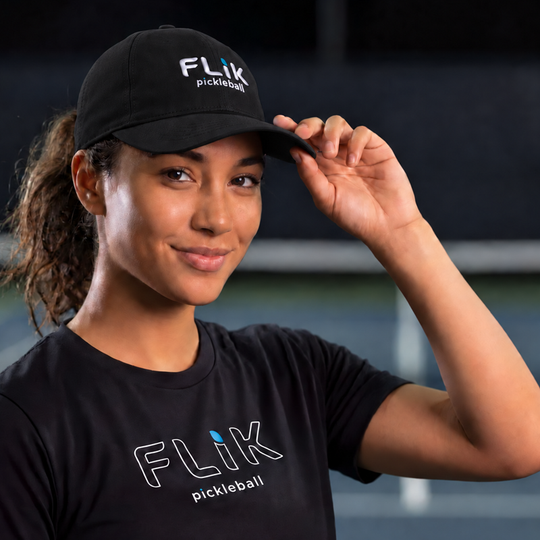 A woman on a pickleball court wearing a black FliK pickleball cap and a black FliK pickleball t-shirt, adjusting her cap with one hand.