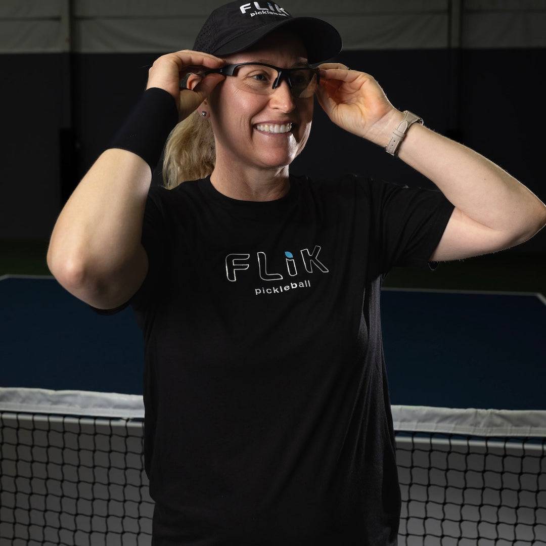 Person wearing a black 'Flik Pickleball' shirt and cap, standing in front of a pickleball net.