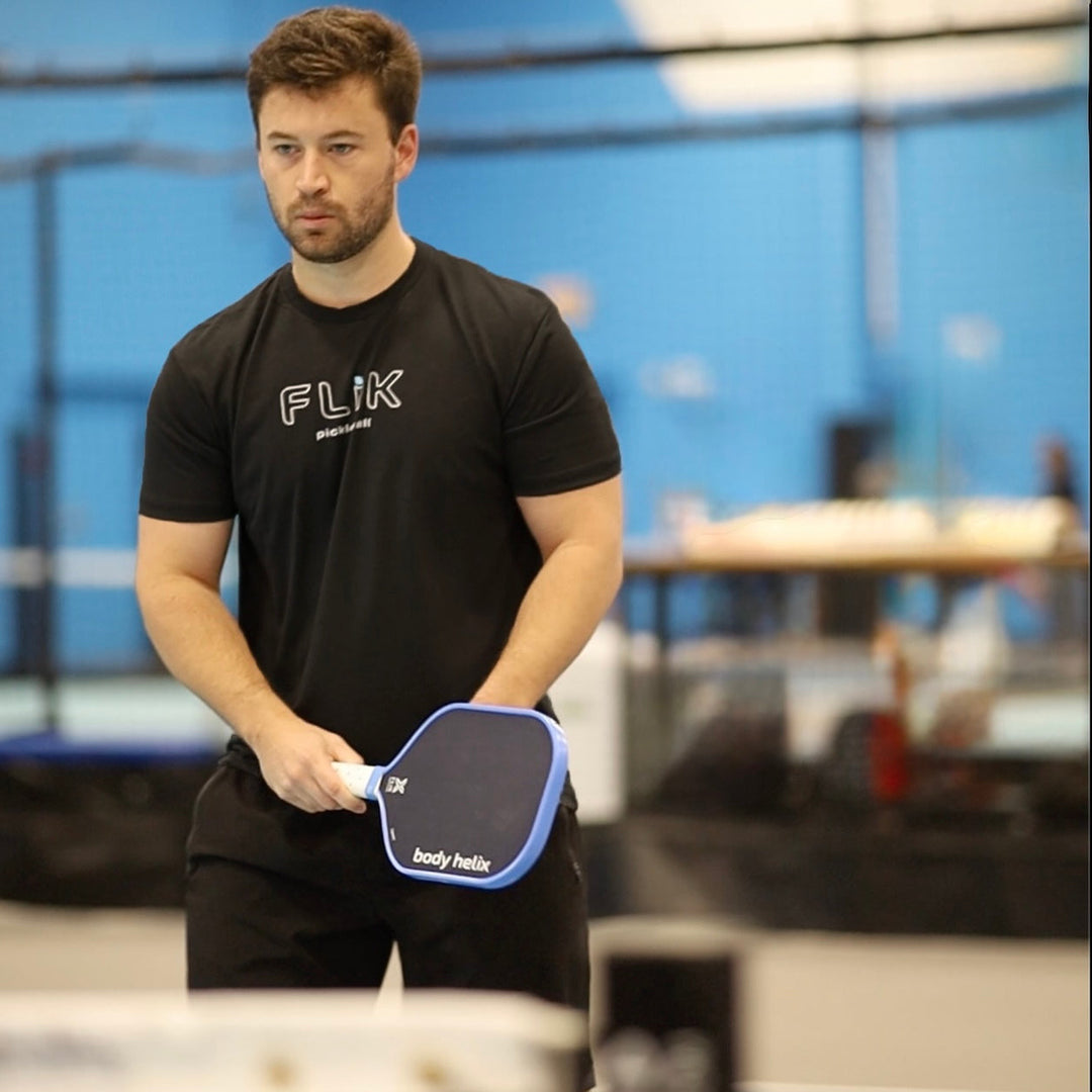 Man holding a FLiK pickleball paddle with a blurred indoor sports facility background