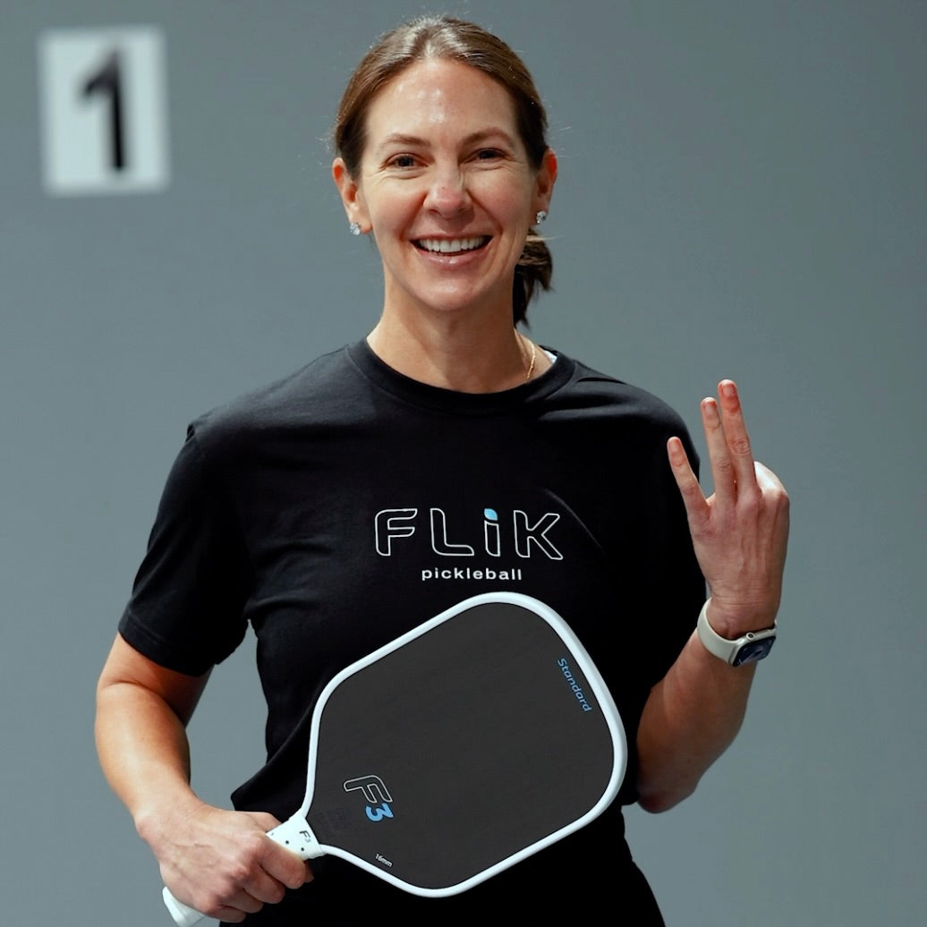 Woman wearing a black 'Flik pickleball' shirt holding a pickleball paddle against a gray background.