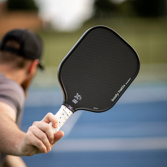 Person holding an X5 pickleball paddle with 'body helix' branding on a blurred pickleball court